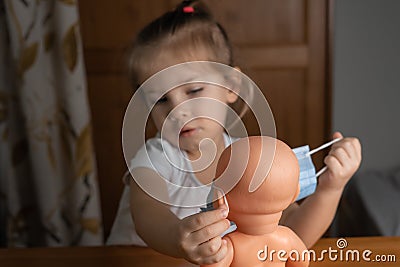 a little girl plays with a baby doll at home, puts a medical mask on a doll. focus on the doll, child in blur Stock Photo