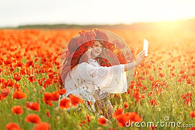 Little girl with redhead mother in white dresses and wreathes make selfie with bouquet of poppies on poppy field at summer sunset Stock Photo