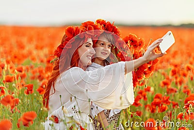 Little girl with redhead mother in white dresses and wreathes make selfie with bouquet of poppies on poppy field at summer sunset Stock Photo