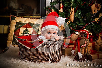 Little girl in Santa hat sits in basket against the background of fireplace and Christmas tree and close looks at the camera Stock Photo