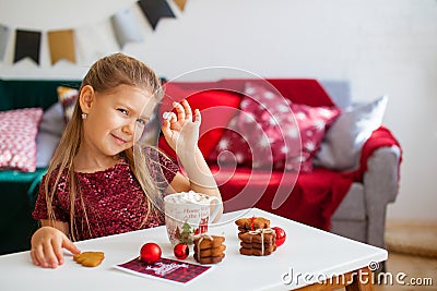 Little girl in red dress eating Christmas cookies with cacao in cup, red Chirstmas decorations around Stock Photo