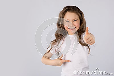 Little girl showing thumbs up gesture in a white T-shirt on white background. Stock Photo