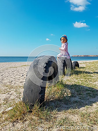 A little girl sits near the sea on a tire buried in the ground until half Stock Photo