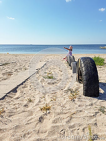 A little girl sits near the sea on a tire buried in the ground until half Stock Photo