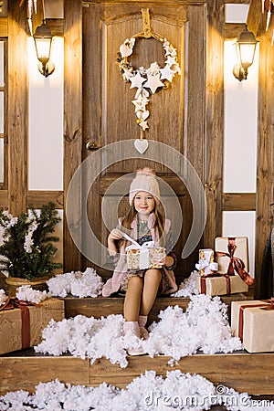 A little girl sits on the porch of a house decorated for Christmas and New Year. She holds a gift box in her hands, opens it by th Stock Photo