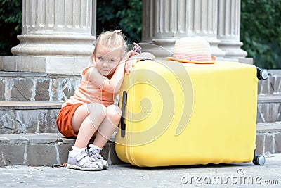 A little girl sits on the steps and leans on a big yellow suitcase. A beautiful child is looking at the camera. Stock Photo