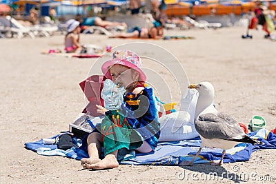 Little girl sits on towel on the beach, eats Honey Baklava and avoid the seagull which watches the action and wants to steal a pie Stock Photo