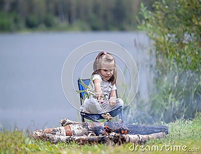 Little girl sitting near campfire. Girl roasting bread over campfire Stock Photo