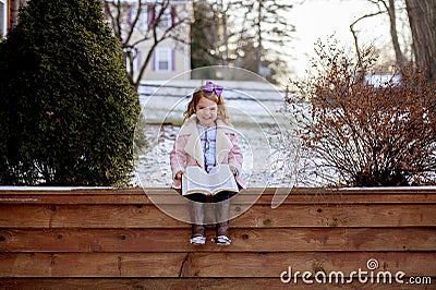 Little girl sitting on wooden planks and reading the bible in a garden covered in the snow Stock Photo