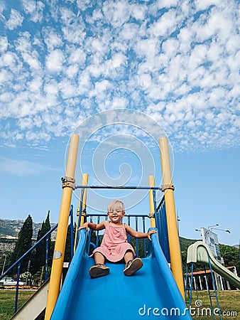 A little girl is sliding down a children`s slide against the background of a blue sky with white clouds. Stock Photo