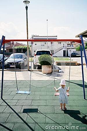 Little girl stands near the swing on the playground and holds on to the chains Stock Photo