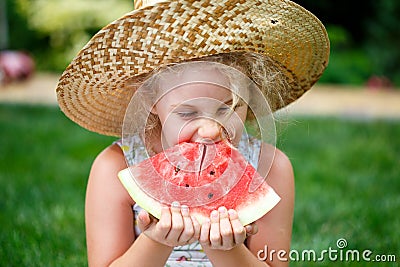 Little girl in straw hat with big slice of watermelon sitting on green grass in summer park. Stock Photo