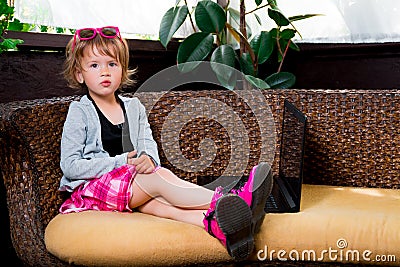 Little girl using laptop. Child in pink skirt and boots, sunglasses, grey top sitting at the sofa, playing . Looking the camera Stock Photo