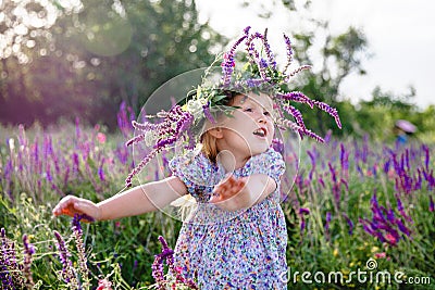 A little happy blonde girl in a sage wreath and a colorful dress in a summer blooming field Stock Photo