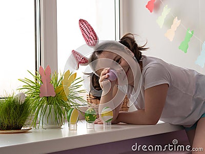 A little happy girl with rabbit ears in a white T-shirt near the window holds a painted Easter egg in her hand, puts it to her eye Stock Photo