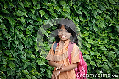 Little Indonesian schoolgirl walks with a school bag against a background of a wall with plants Editorial Stock Photo
