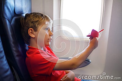 Little kid boy playing with red paper plane during flight on airplane. Child sitting inside aircraft by a window. Family Stock Photo