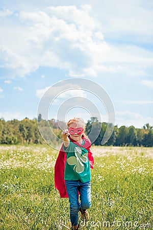 little kid in red superhero cape and mask with outstretched arm running in meadow Stock Photo