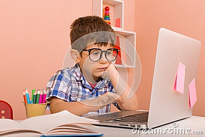 Unhappy little kid sit and study at white desk with pc laptop isolated on pastel pink background. Stock Photo