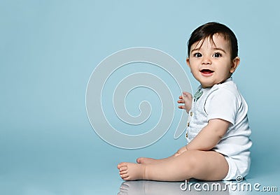 Little kid in white bodysuit as vest with bow-tie, barefoot. He smiling, sitting on the floor against blue background. Close up Stock Photo