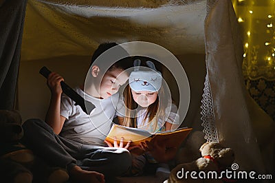 Little kids involving in reading amazing book. They lying in nice toy tent in playroom. Boy holding flashlight in hand. Stock Photo