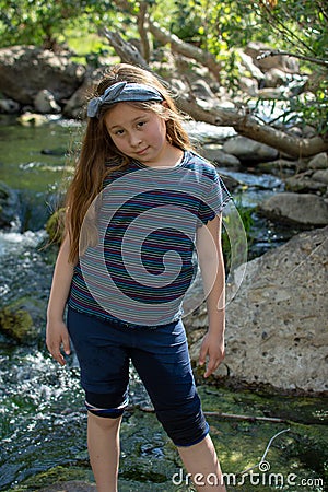 Little latina girl smiling coyly and looking down while standing in front of a stream and woods in the shade Stock Photo