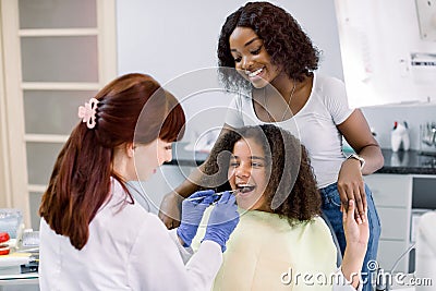 Little mixed raced school girl with curly hair, visiting dentist for checkup or caries treatment, sitting in dental Stock Photo