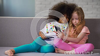 Little multi-ethnic sisters on couch, hugging and taking photos on smartphone Stock Photo