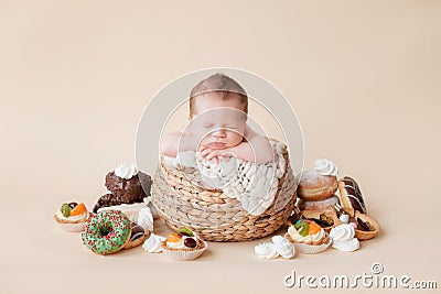 A little newborn baby sleeps in a straw basket on a beige background, there are many donuts, cakes and sweets Stock Photo