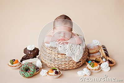 A little newborn baby sleeps in a straw basket on a beige background, there are many donuts, cakes and sweets Stock Photo
