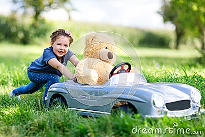Little preschool kid girl driving big toy car and having fun with playing with big plush toy bear Stock Photo