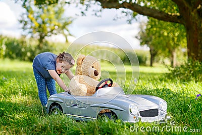 Little preschool kid girl driving big toy car and having fun with playing with big plush toy bea Stock Photo