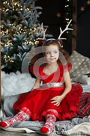 Little pretty curly smiling girl sitting nearly Christmas tree with Christmas decorations and presents. Child in red dress and soc Stock Photo