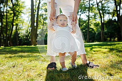Little pretty girl in white dress holds mom by the hands and tries to make her first steps. Stock Photo