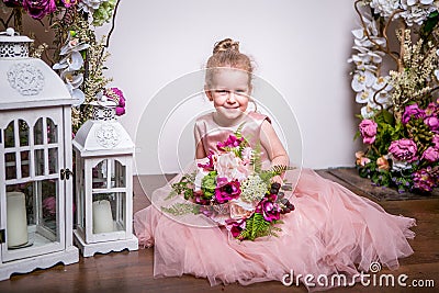 A little princess in a beautiful pink dress sits on the floor near flower stands and lanterns, holds a bouquet of peonies, magnoli Stock Photo