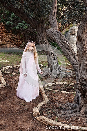 A little princess of spirits and fairies stands in the enchanted mysterious magic forest in a wedding dress with a veil and crown Stock Photo