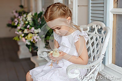 The little red-haired girl with pigtails holding a yellow chicken on a background of white houses and flowers Stock Photo