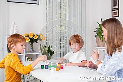 Little redhead boys with a nanny or mother or teacher sit at the table and paint with colors Stock Photo