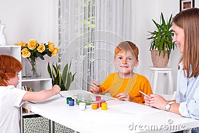 Little redhead boys with a nanny or mother or teacher sit at the table and paint with colors Stock Photo