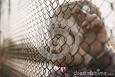 A little refugee girl with a sad look behind a metal fence. The social problem of refugees and internally displaced persons Stock Photo