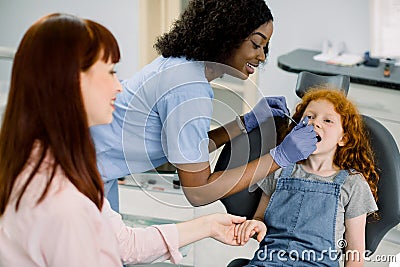Little school girl with red curly hair, visiting dentist for checkup or caries treatment, sitting in dental chair and Stock Photo