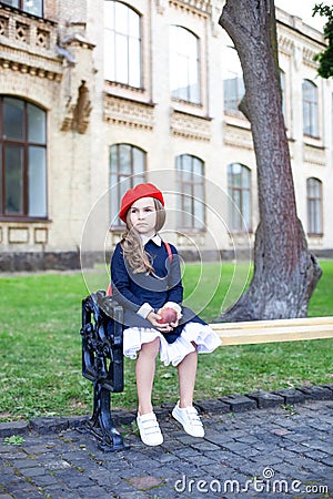 Little schoolgirl in a red beret and dress with lunch near the school. preschool child with an apple and a backpack on his first d Stock Photo