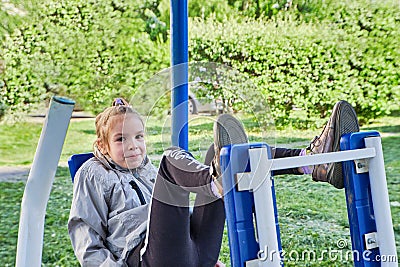 A little sporty girl in a sports uniform is engaged in the exercise machines on the playground for fitness on the street Stock Photo