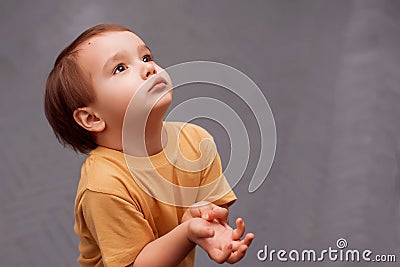 Little toddler boy in yellow shirt standing and looking up asking for something. The kid is holding hands raised to chest like he Stock Photo