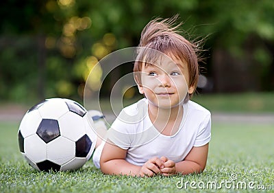 Little toddler child laying on belly on football field near soccer ball and dreaming. Wind is waving hair of kid. Future sportsman Stock Photo