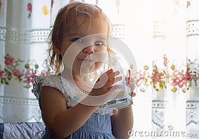 Little toddler girl child drinks from a glass goat or cow milk, natural rustic. The happy kid smiles and looks pleased Stock Photo