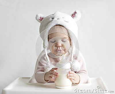 Little toddler girl in a warm fluffy hat drinks milk from a bottle while sitting. Half-length portrait. White gray Stock Photo