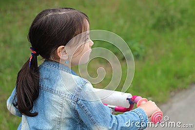 A little white girl with dark hair and blue earrings tied in a ponytail rides a pink bicycle. Beautiful child in denim jacket on Stock Photo