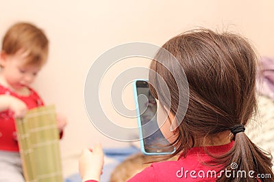 A little white girl with a ponytail takes a picture of her sister on a cell phone against the background of a regular apartment Stock Photo