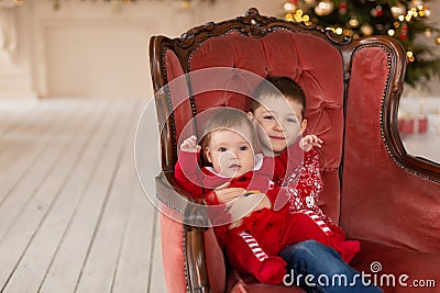 Litttle brother hugs his little sister in red retro chair near Christmas Tree. Enjoying a love hug, people`s holidays. Stock Photo